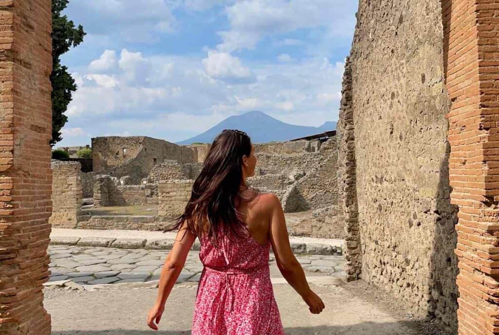 woman with long brown hair and red dress in the ruins of Pompeii in front of mount Vesuvius in Italy