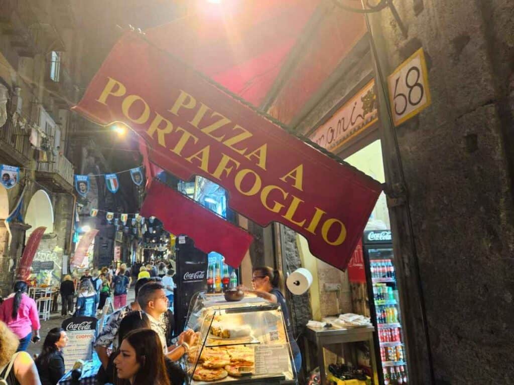 View of a busy street in the old town of Naples at night with a pizza a portafoglio sign (folded pizza).
